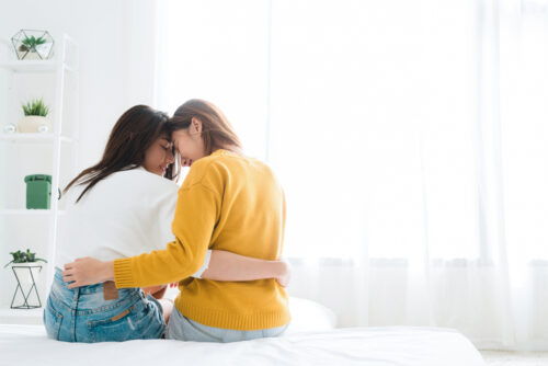 Back view of lesbian happy couple waking up in morning, sitting on bed, stretching in cozy bedroom, looking through window. 